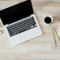 Offener Laptop, Kaffeetasse und gelber Notizblock mit Stiften auf hellem Holzschreibtisch