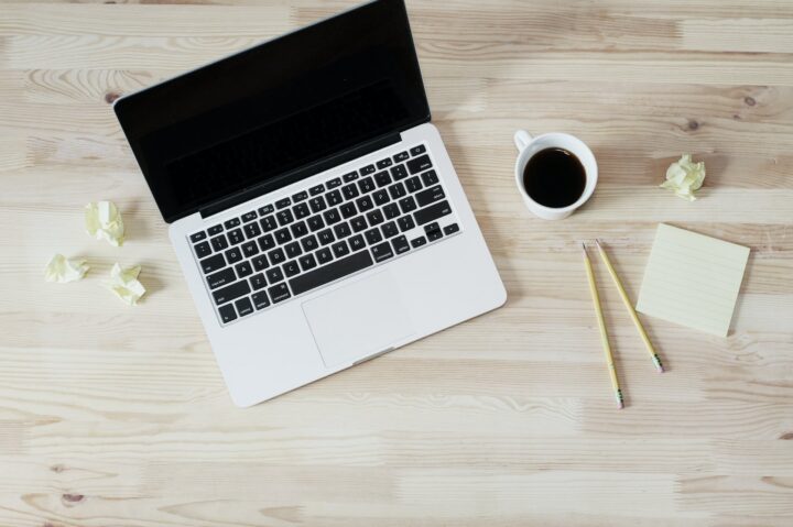 Offener Laptop, Kaffeetasse und gelber Notizblock mit Stiften auf hellem Holzschreibtisch