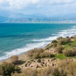 Mittelmeerküste mit antiken Ruinen auf einem Gras hang und türkisem Meer vor Bergkulisse