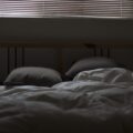 Ungemachtes Bett mit weißer Bettdecke und zwei Kissen in einem abgedunkelten Schlafzimmer, darüber eine geschlossene Jalousie