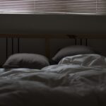Ungemachtes Bett mit weißer Bettdecke und zwei Kissen in einem abgedunkelten Schlafzimmer, darüber eine geschlossene Jalousie