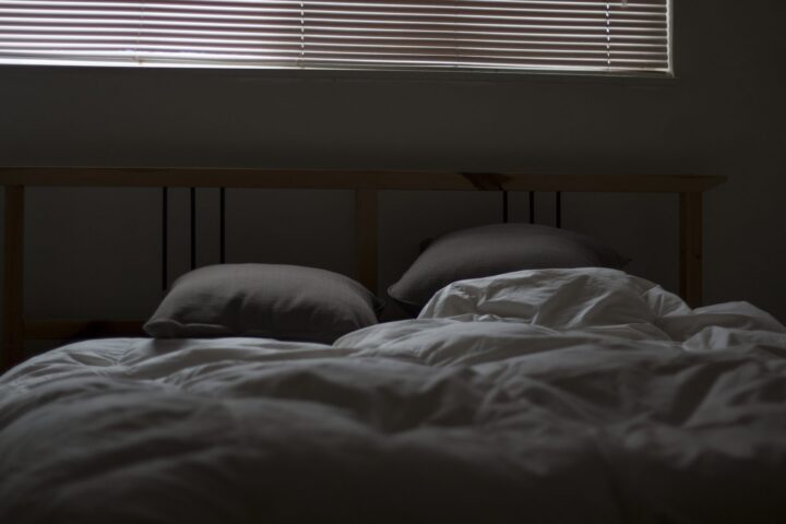 Ungemachtes Bett mit weißer Bettdecke und zwei Kissen in einem abgedunkelten Schlafzimmer, darüber eine geschlossene Jalousie
